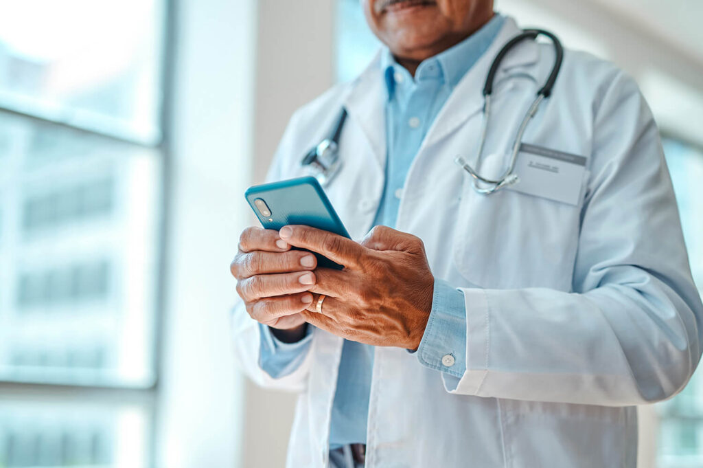 A doctor wearing a white lab coat and a stethoscope around the neck is holding and looking at a blue smartphone. The doctor's face is partially visible, and the background shows large windows with blurred outdoor scenery